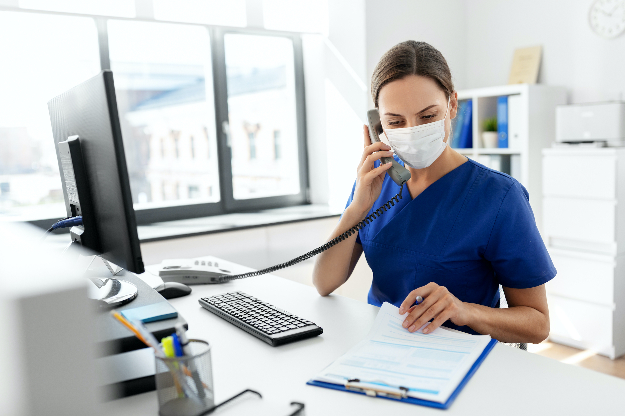 medicine, people and healthcare concept - female doctor or nurse wearing face protective medical mask for protection from virus disease with computer and clipboard calling on phone at hospital